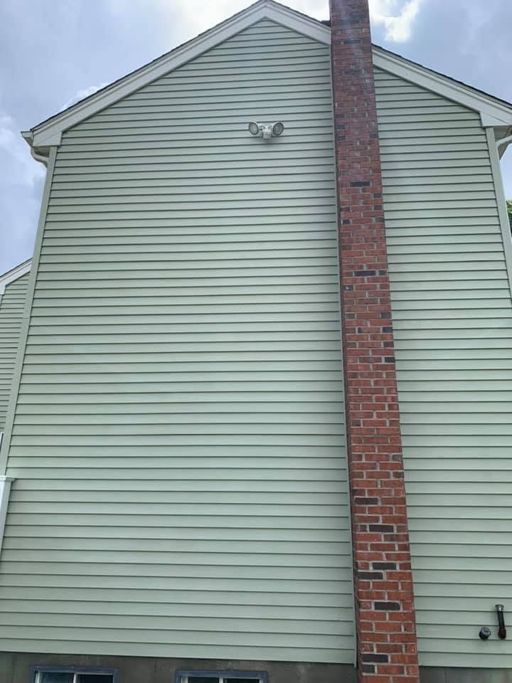 Side of a light green house with a red brick chimney and gabled roof under a cloudy sky