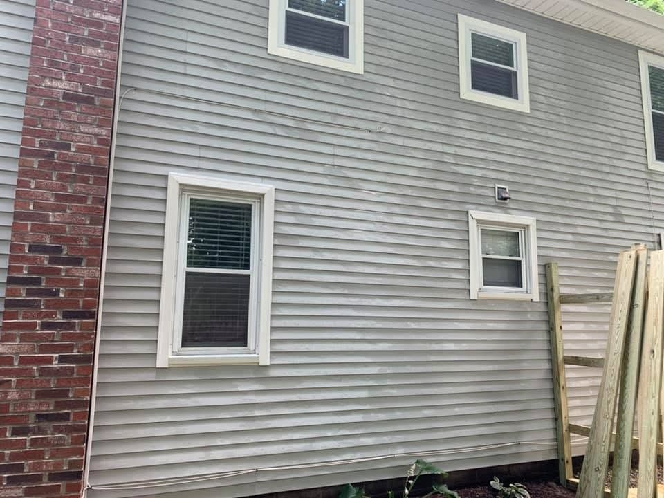 Gray house siding with two windows, a brick corner, and wooden boards leaning on the right.