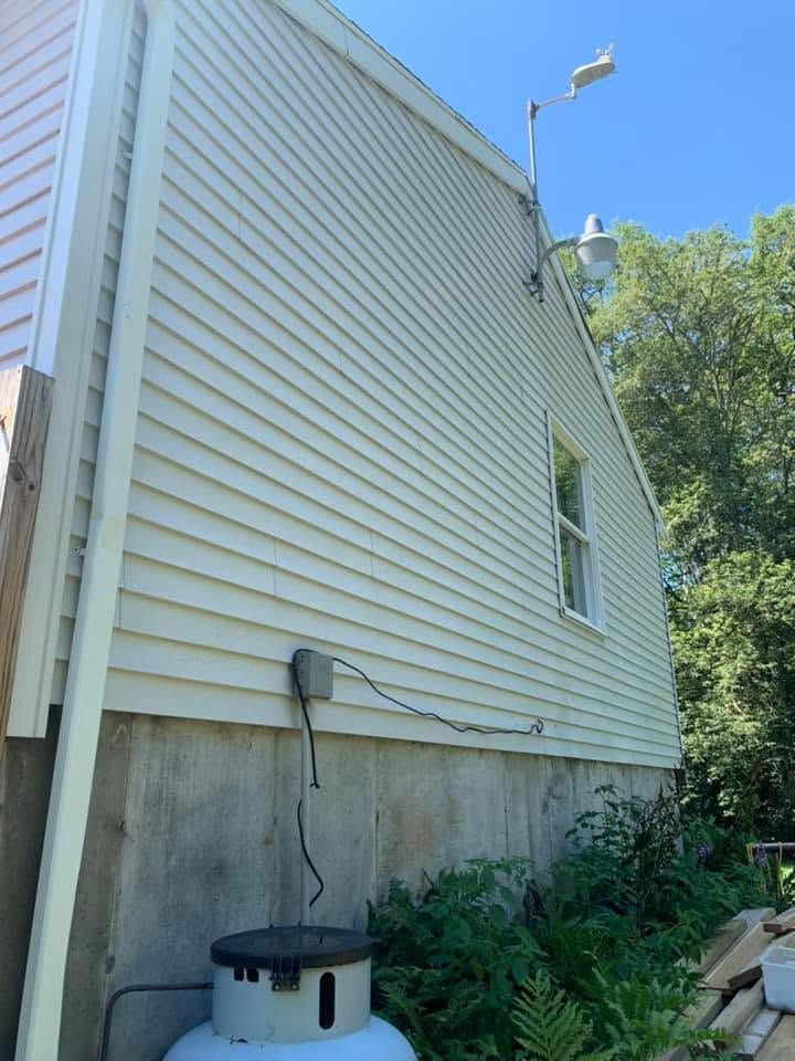 Side view of a house with beige siding, concrete foundation, and overgrown plants beside it under a blue sky