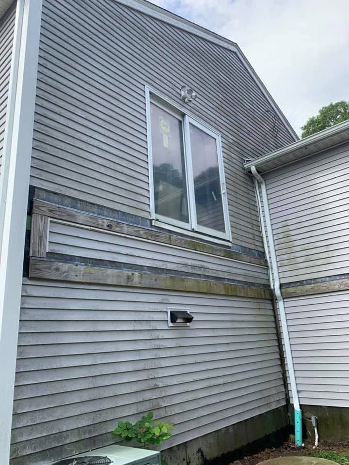 Side of a gray house with weathered siding, a second-story window, and a downspout by a small plant.