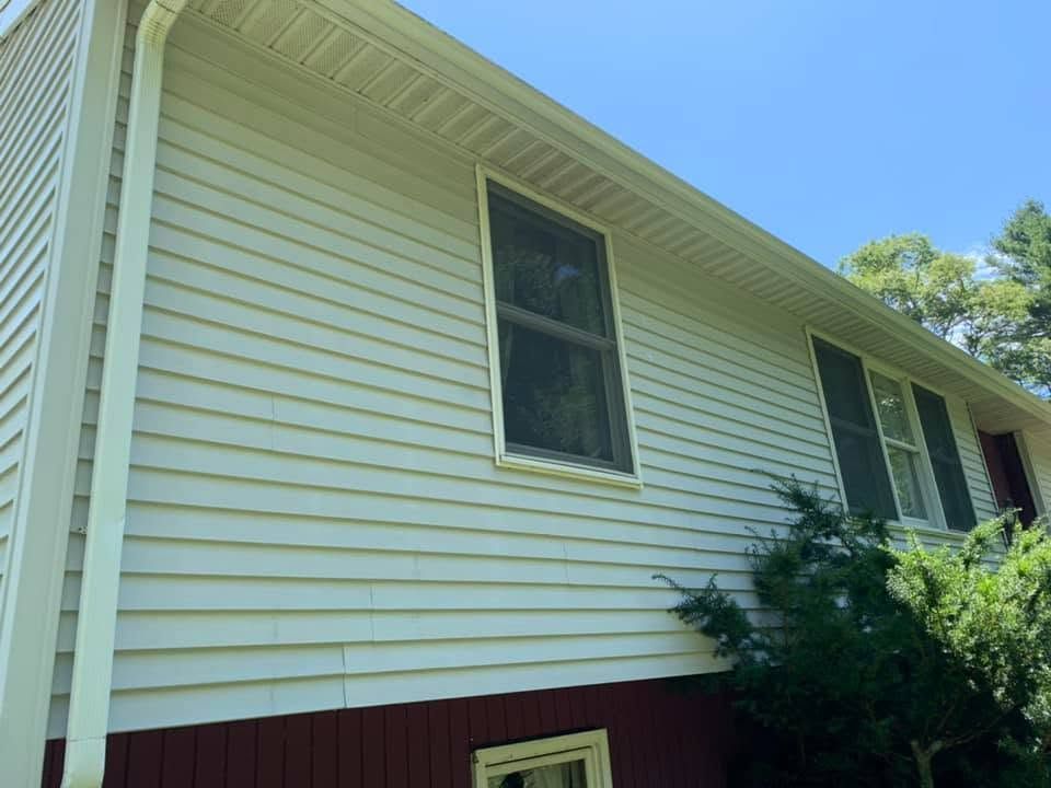 Side view of a white house with beige siding, a dark window, and bushes against a blue sky.