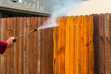 Person pressure-washing a wooden fence, spraying orange-tinted grime off the panels.