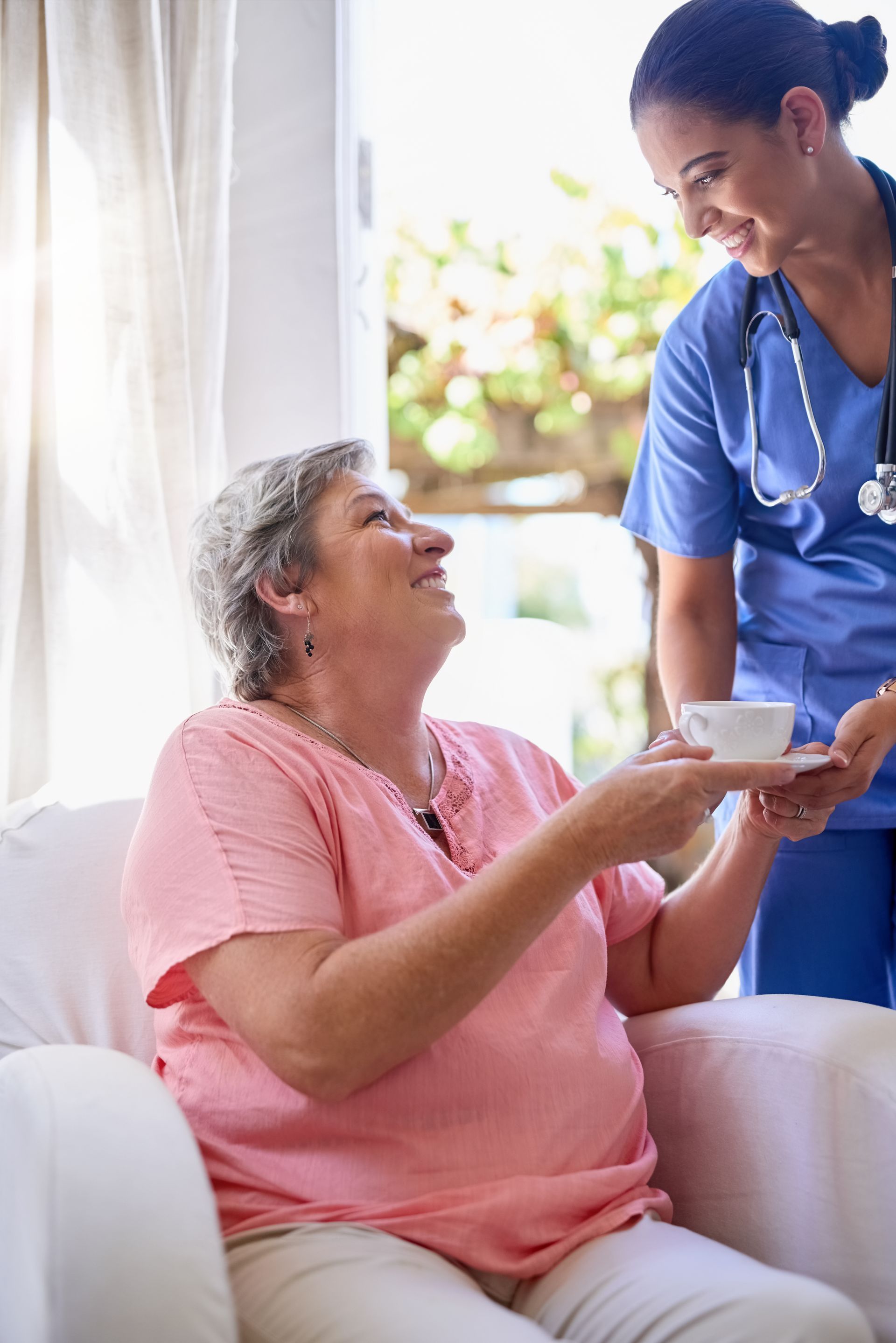 A healthcare worker in blue scrubs hands a cup to a person sitting in a white chair, both smiling warmly.