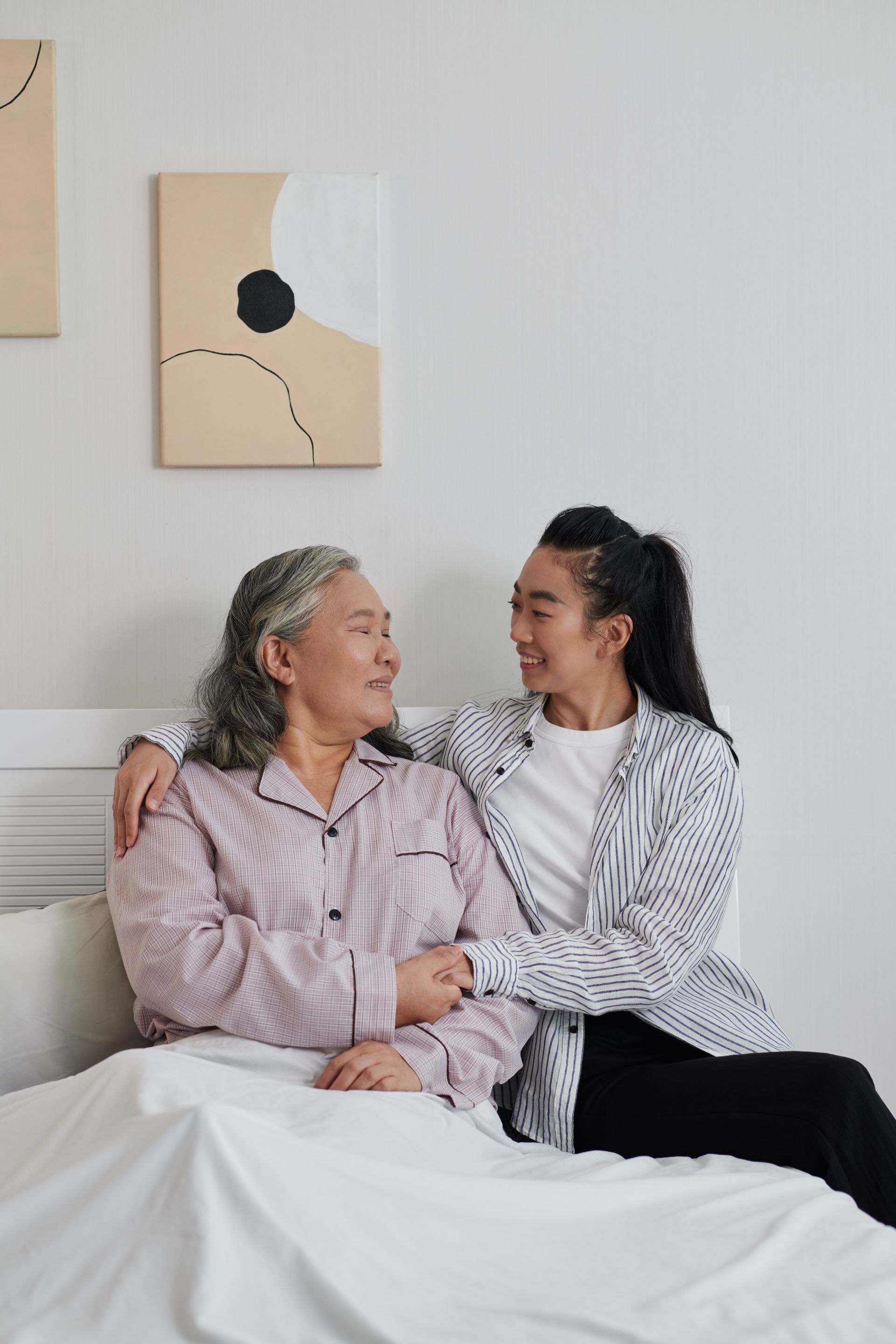 Two people sitting on a bed in a bright room, holding hands and smiling at each other.