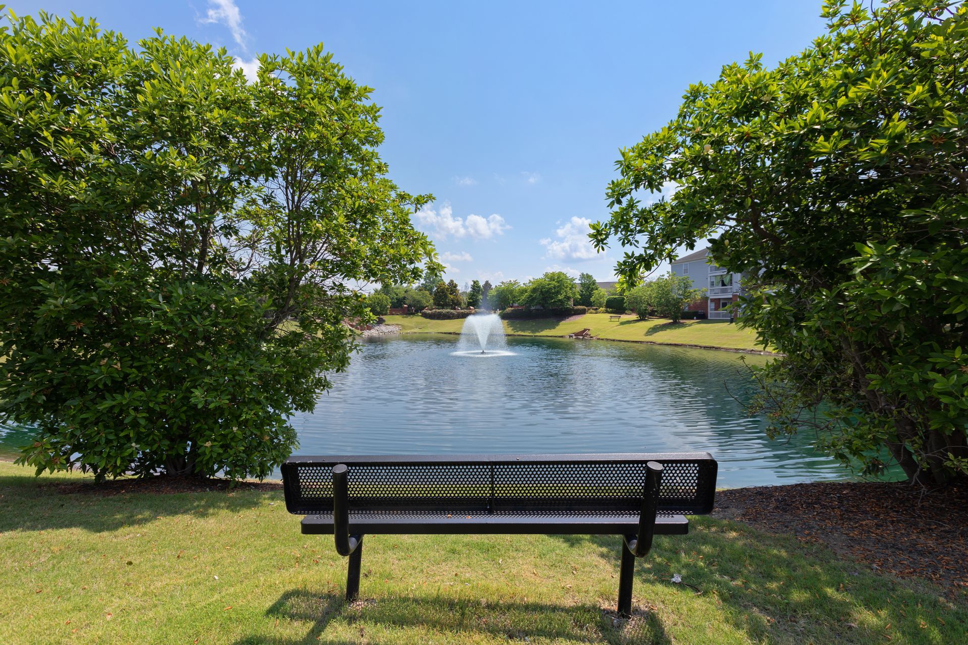 A park bench is sitting in front of a lake with a fountain in the background.