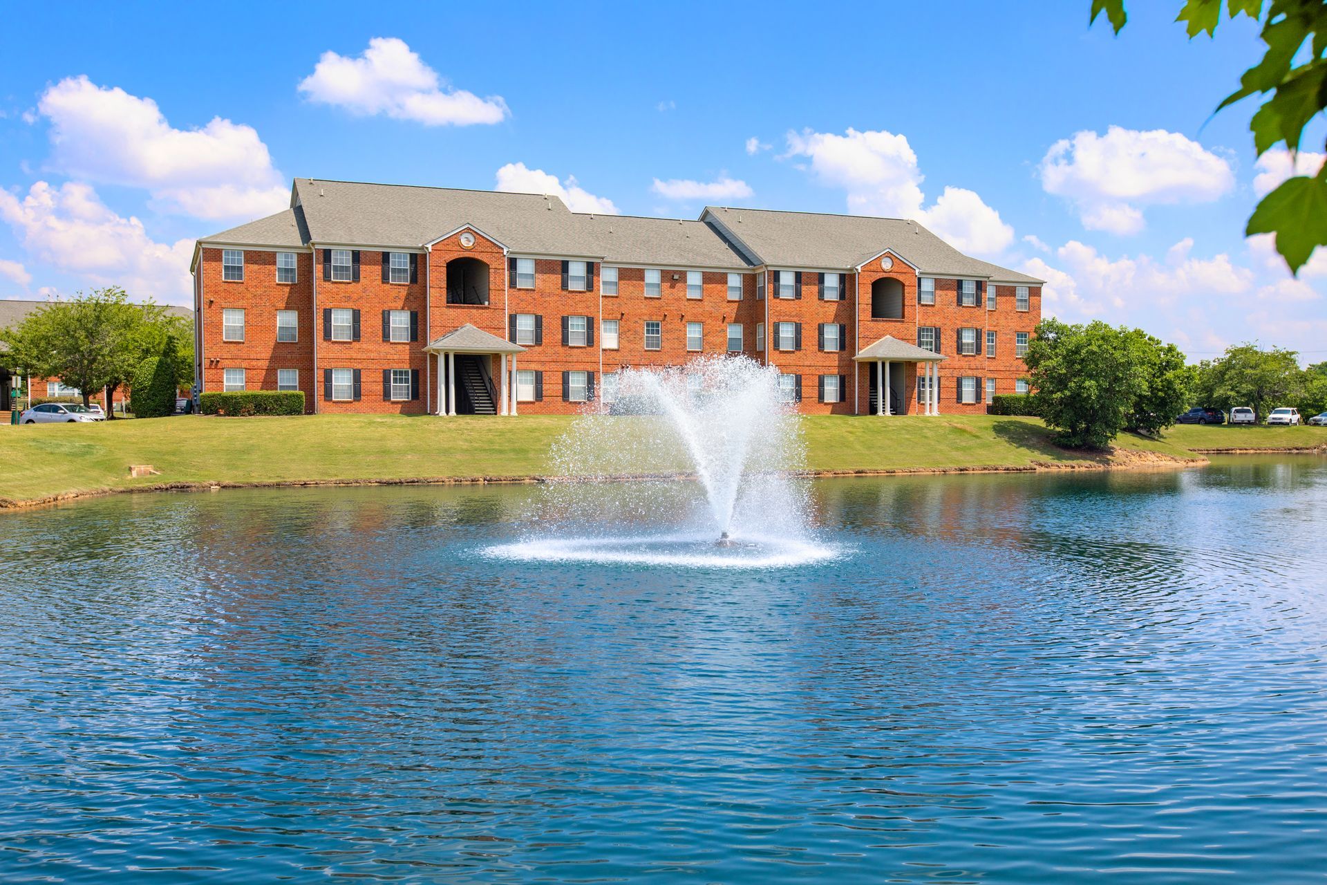 A fountain in the middle of a lake in front of a large brick building.