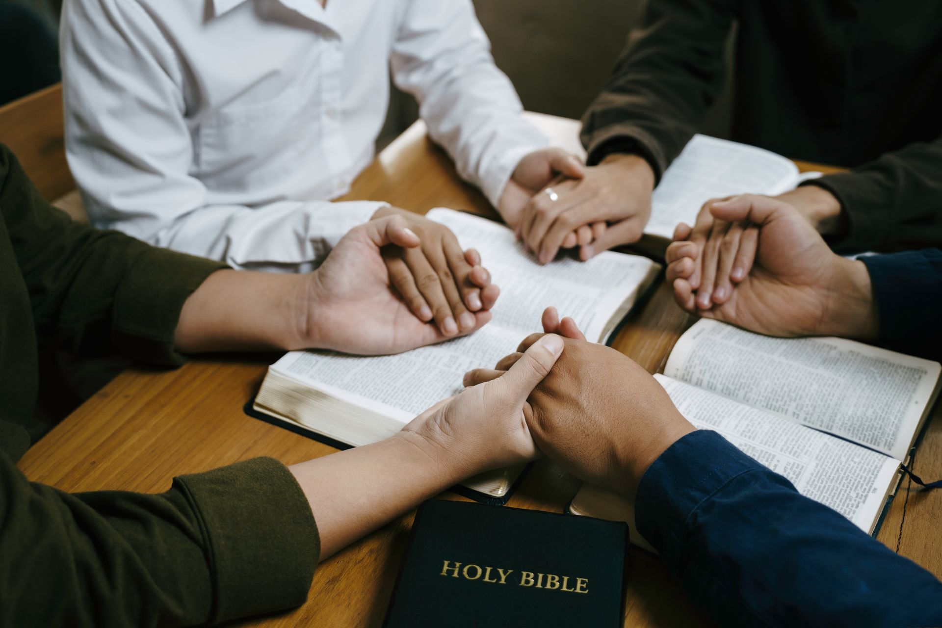People holding hands in prayer around an open Bible on a wooden table