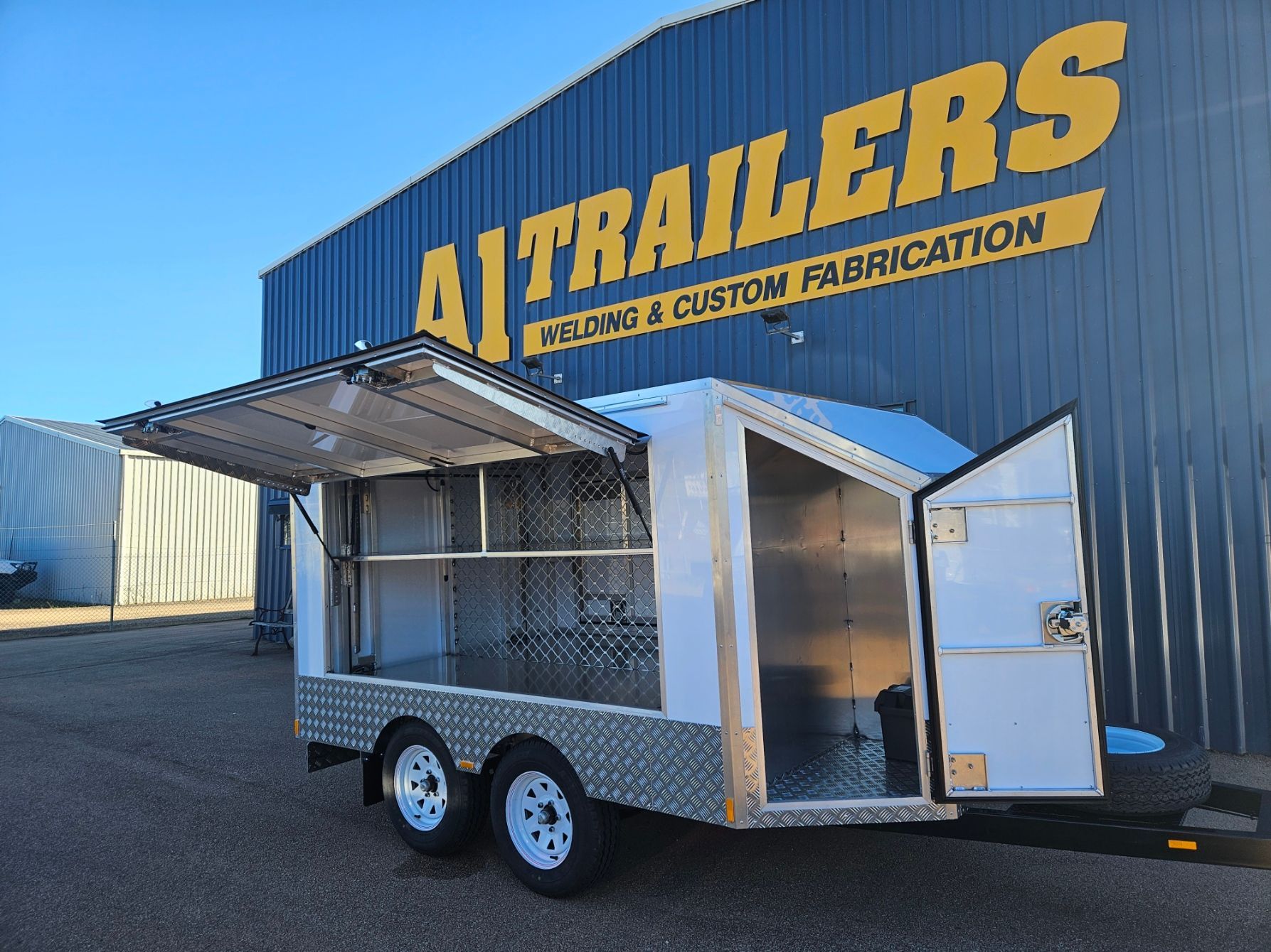 A white custom fabrication trailer with doors open, in front of a blue building with yellow lettering 
