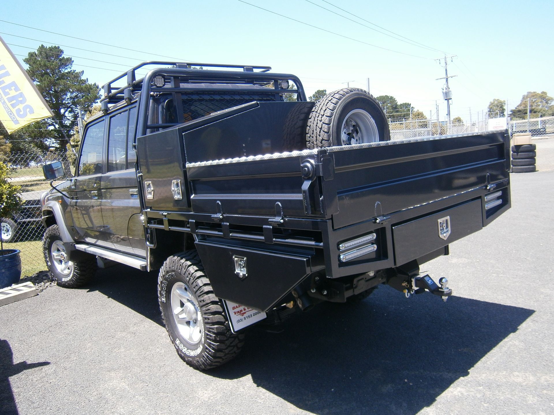 Black utility truck with a flatbed, spare tire, and storage boxes parked outdoors.