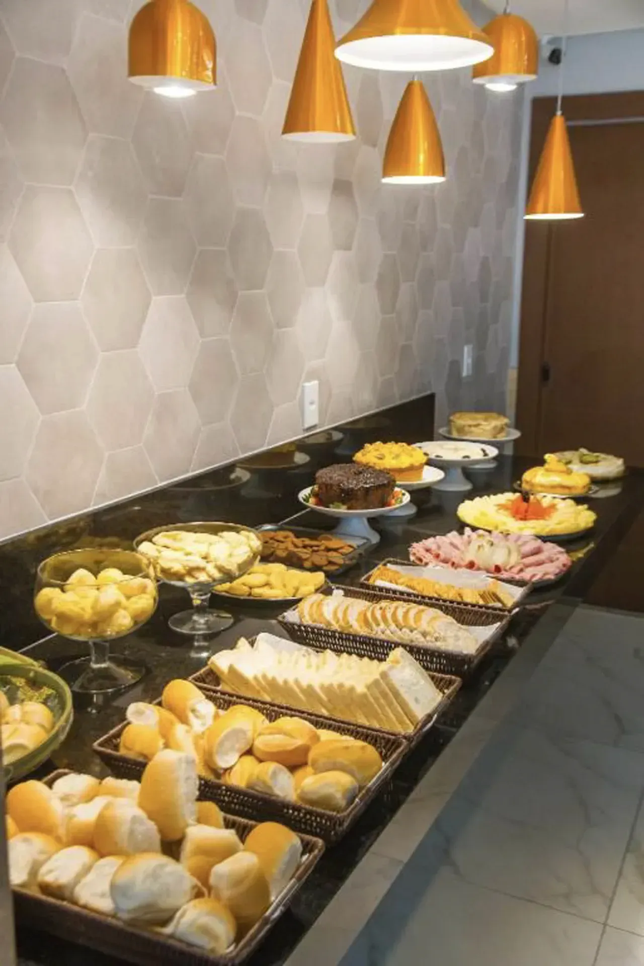Buffet de padaria com uma variedade de pães, doces e recheios em um longo balcão preto sob luzes pendentes.