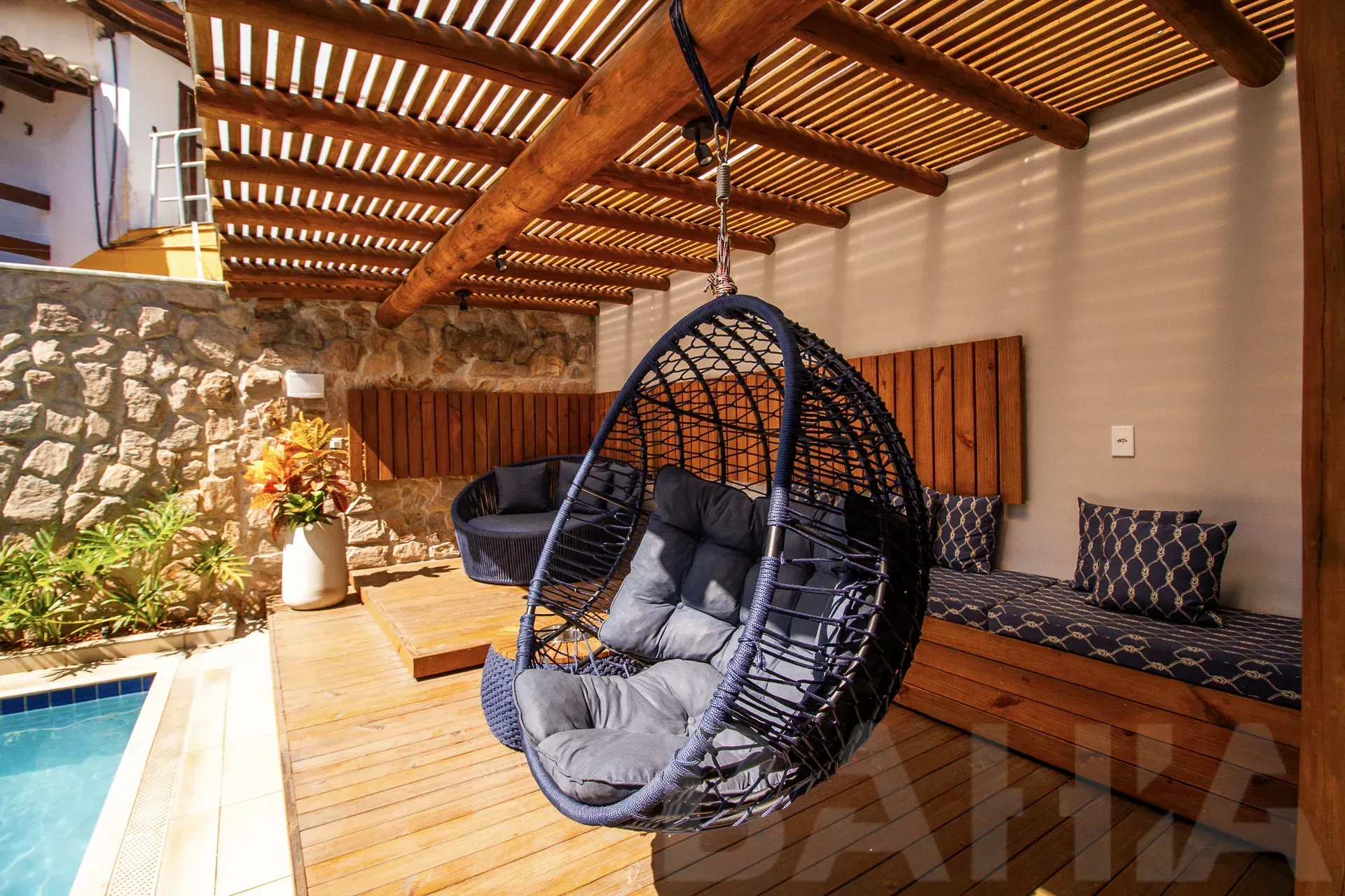 Pátio coberto com cadeira suspensa, sofá e piscina ao lado de um muro de pedra e telhado de ripas de madeira.