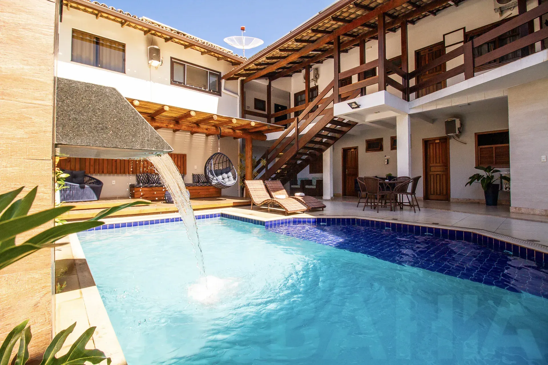 Pátio ensolarado com piscina, fonte, espreguiçadeiras e varandas brancas ao redor.