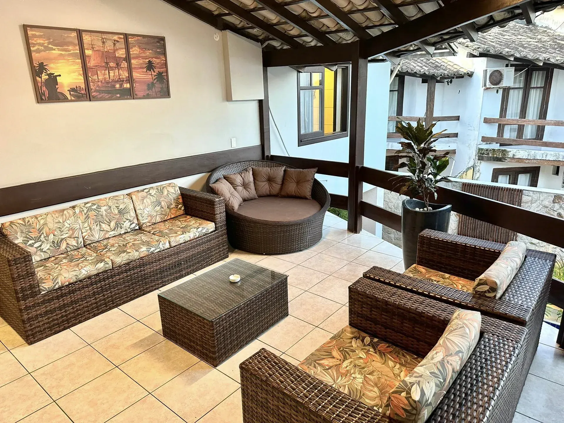 Varanda coberta com assentos de vime, almofadas estampadas e piso de azulejos com vista para um pátio interno.
