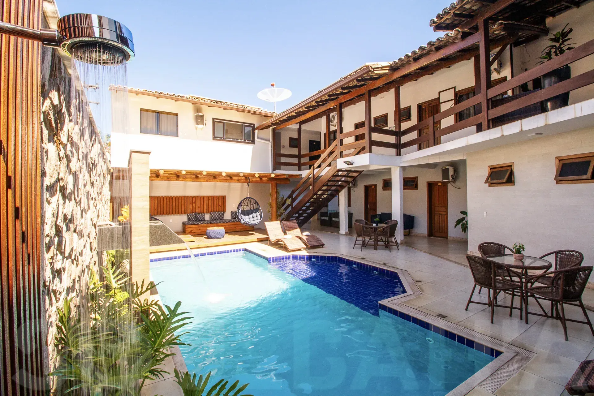 Pátio ensolarado com piscina azul, espreguiçadeiras e área de estar externa ao lado de uma vila branca.