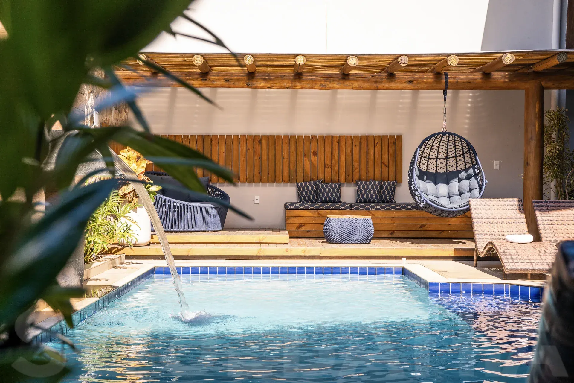 Pátio à beira da piscina com espreguiçadeira almofadada, cadeira suspensa e pérgola de madeira ao lado de uma piscina azul.
