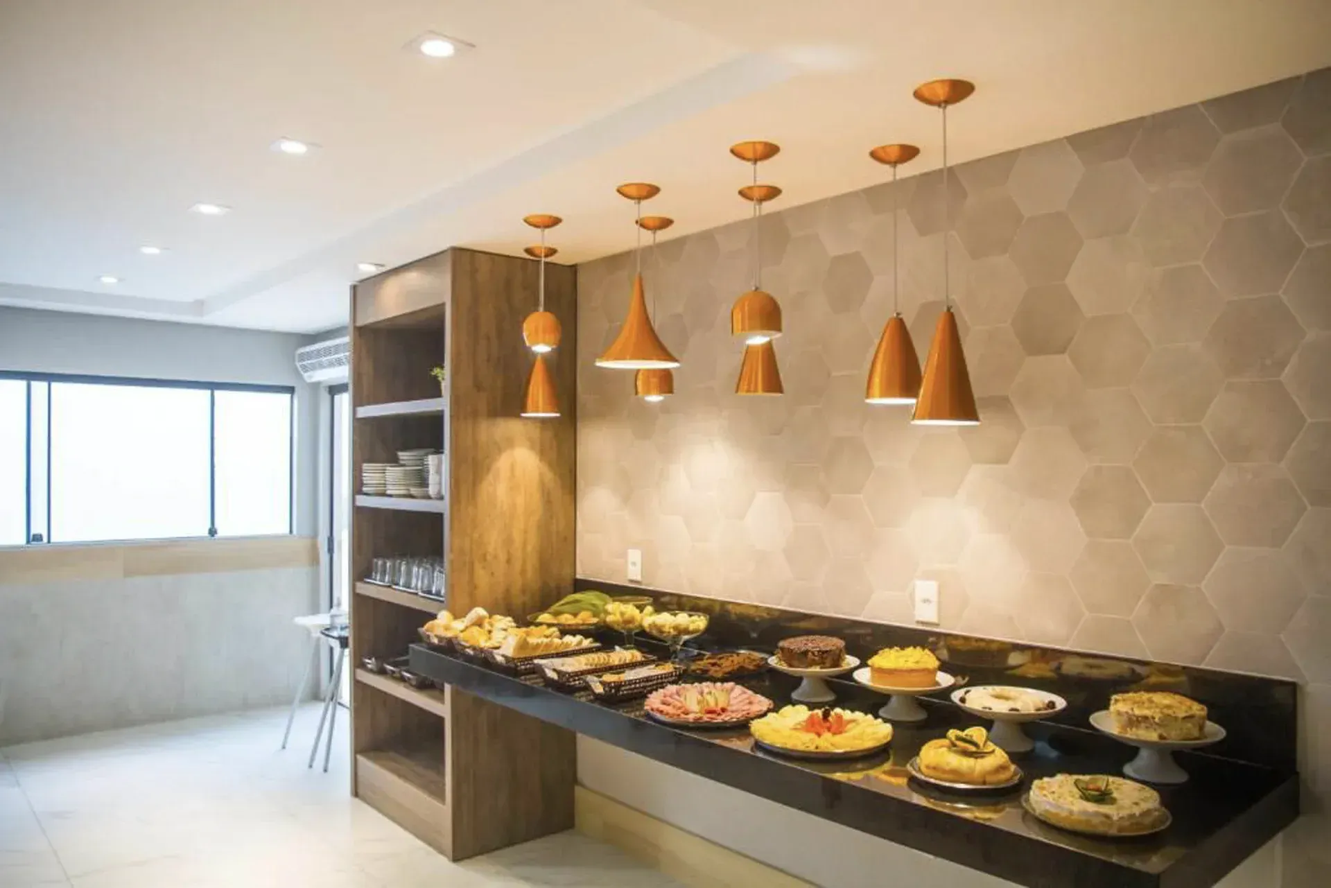 Mesa de buffet moderna com diversos pratos sob luminárias pendentes de cobre em uma sala de jantar iluminada.
