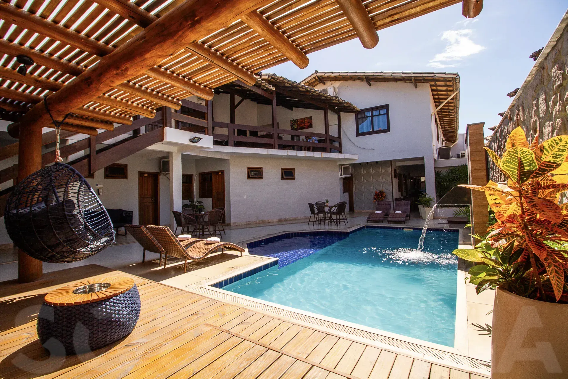 Varanda coberta com cadeira de vime com vista para uma piscina ensolarada no quintal e uma casa de estuque branco.