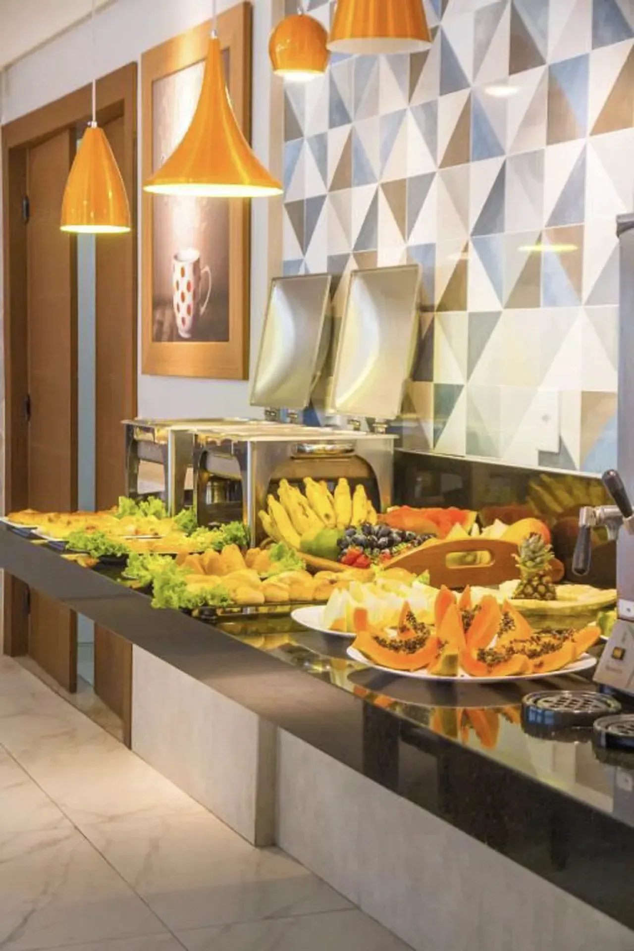 Mesa de buffet com frutas, saladas e doces sob luzes pendentes aconchegantes em uma sala de jantar moderna.