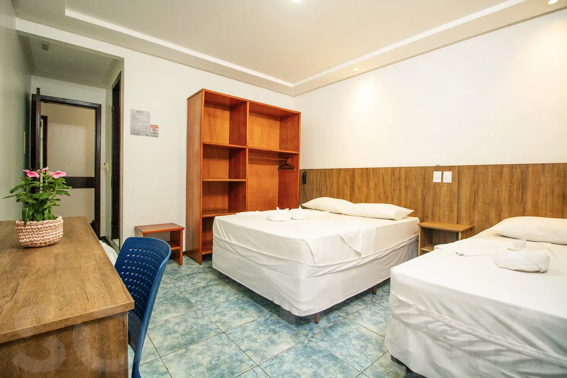 Quarto de hotel com duas camas brancas, prateleiras de madeira, escrivaninha e carpete azul.