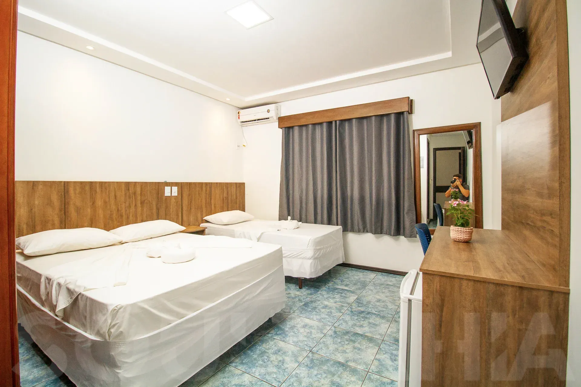 Quarto de hotel impecável com duas camas, painéis de madeira, carpete azul e janela com cortinas cinza.