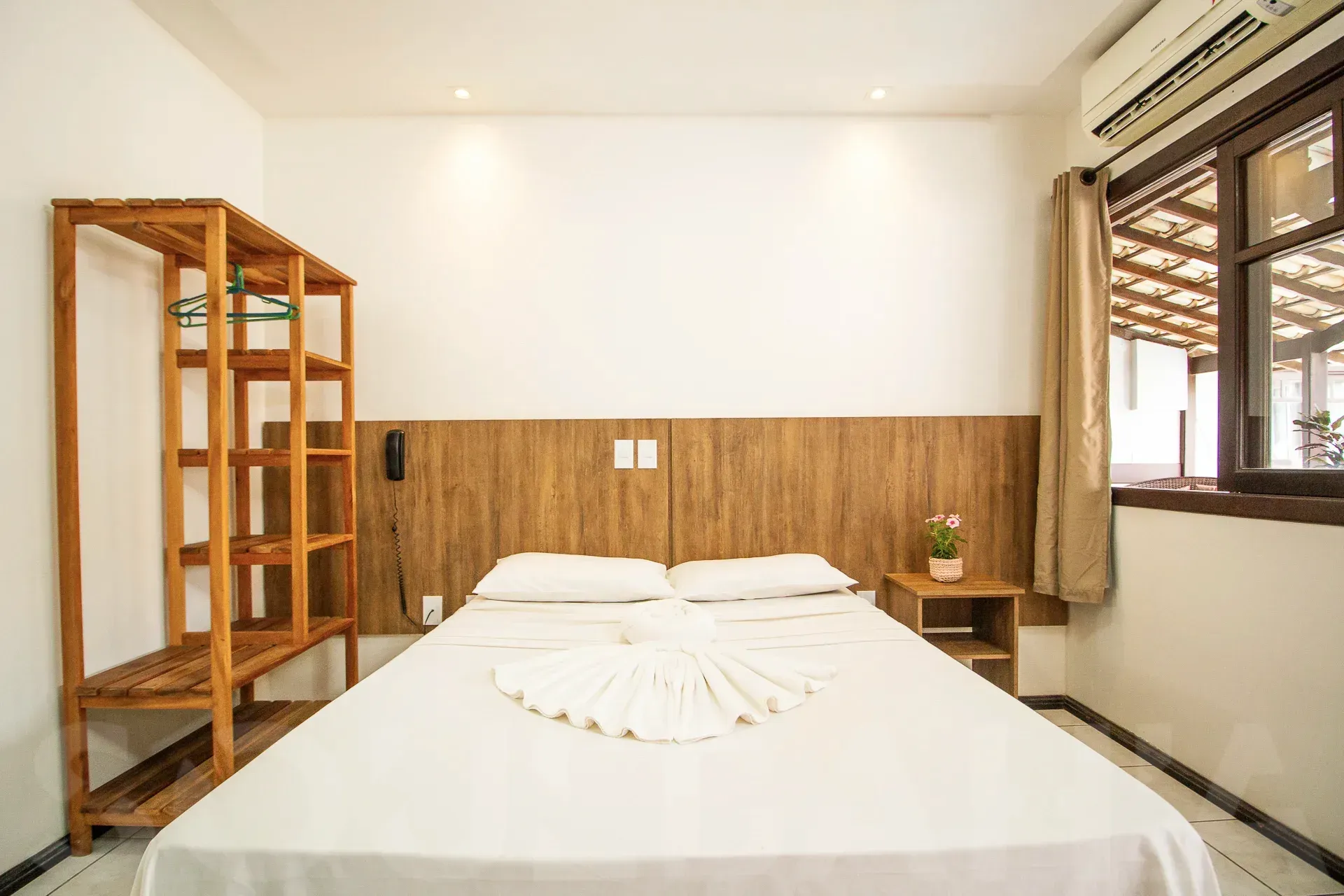 Quarto luminoso com cama, prateleiras de madeira e janela à direita.