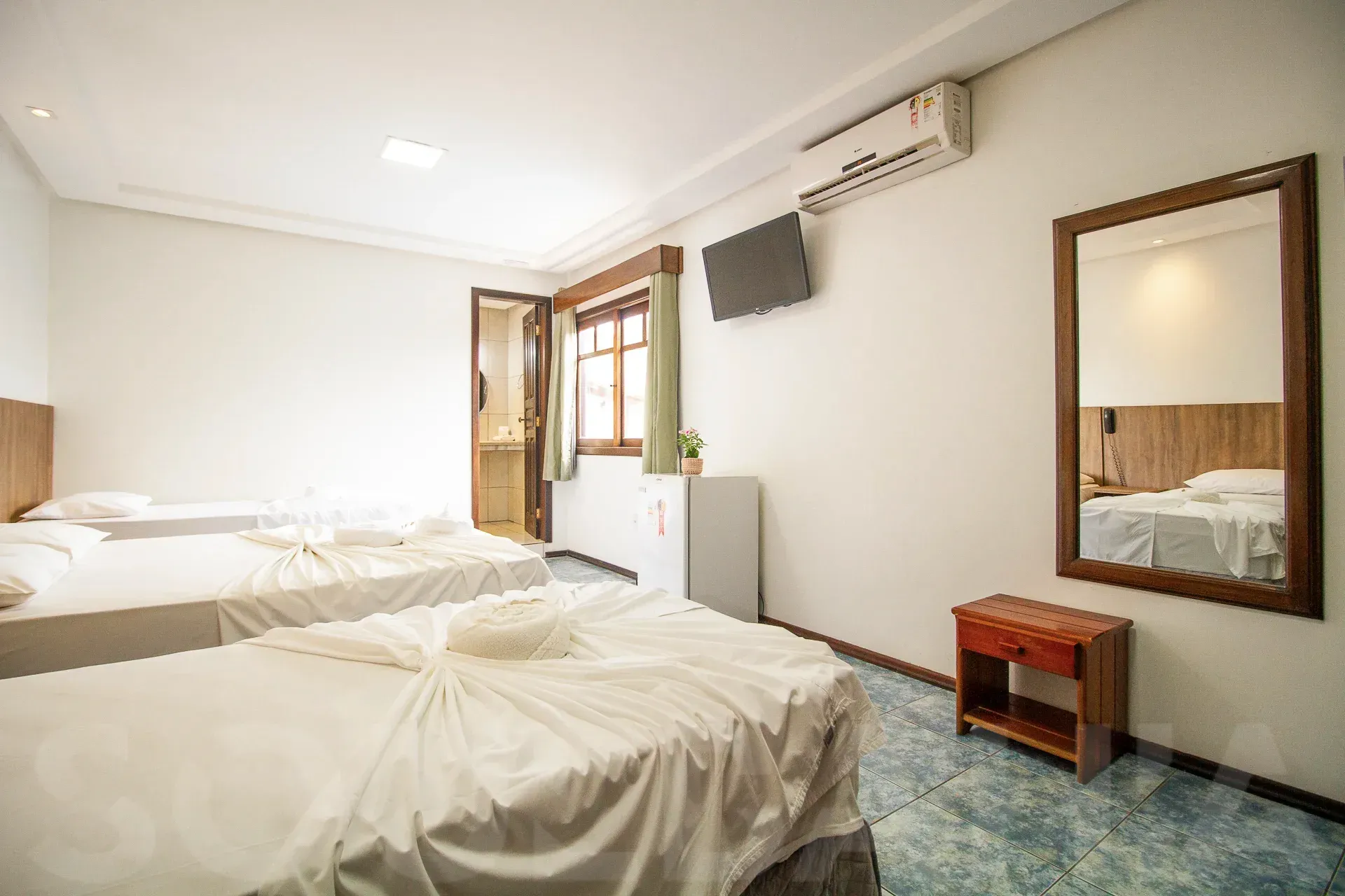 Quarto luminoso com duas camas, janela, espelho de parede e um pequeno banco de madeira sobre um tapete azul.