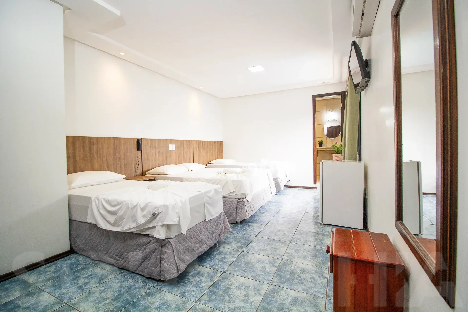 Quarto de hotel pequeno com duas camas de solteiro, paredes brancas e carpete azul, visto da porta.