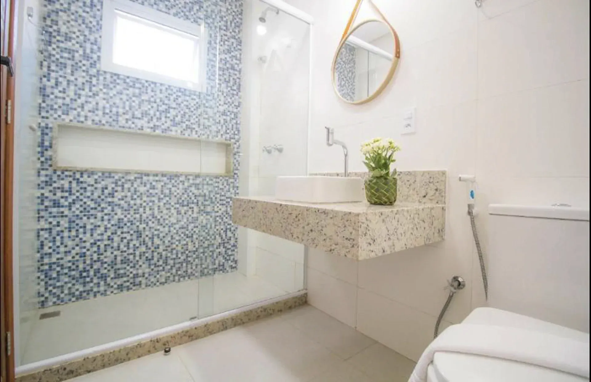 Banheiro moderno e luminoso com azulejos de mosaico azul, pia suspensa, espelho e vaso sanitário.