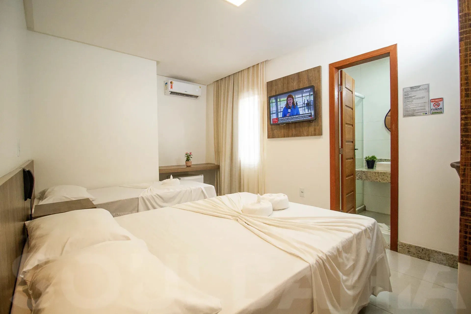 Quarto de hotel luminoso com duas camas brancas, TV de parede, escrivaninha e porta do banheiro aberta à direita.