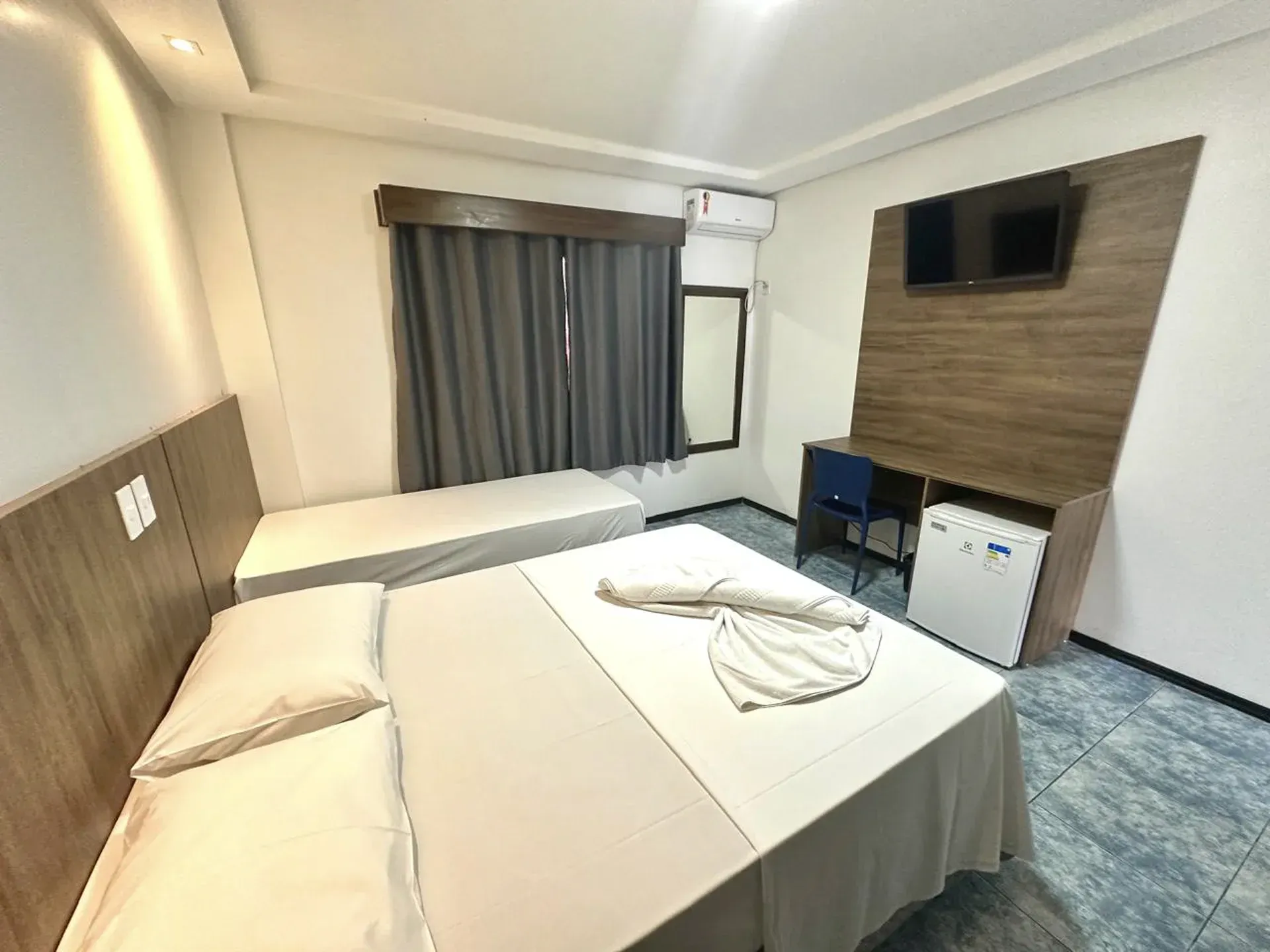 Quarto de hotel pequeno com duas camas de solteiro, cortinas cinza, TV de parede, escrivaninha e frigobar.