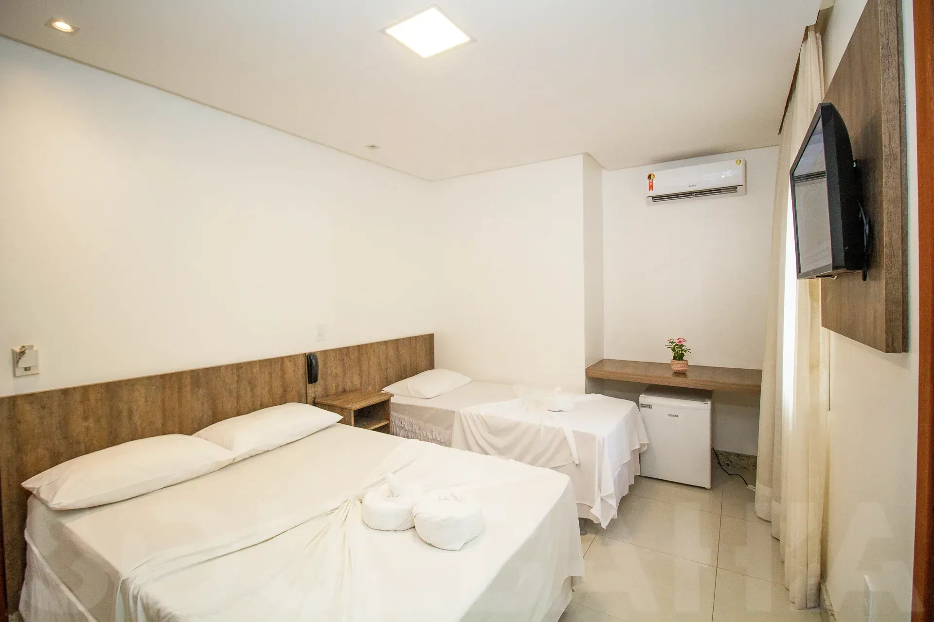 Quarto de hotel luminoso com duas camas, TV de parede, escrivaninha e ar-condicionado.