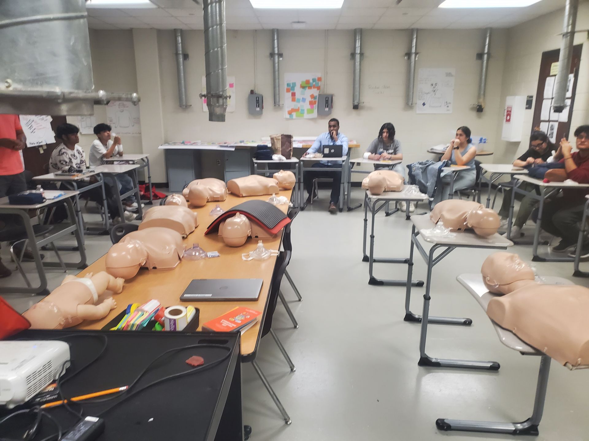 A group of people are sitting at tables in a classroom with mannequins on them.