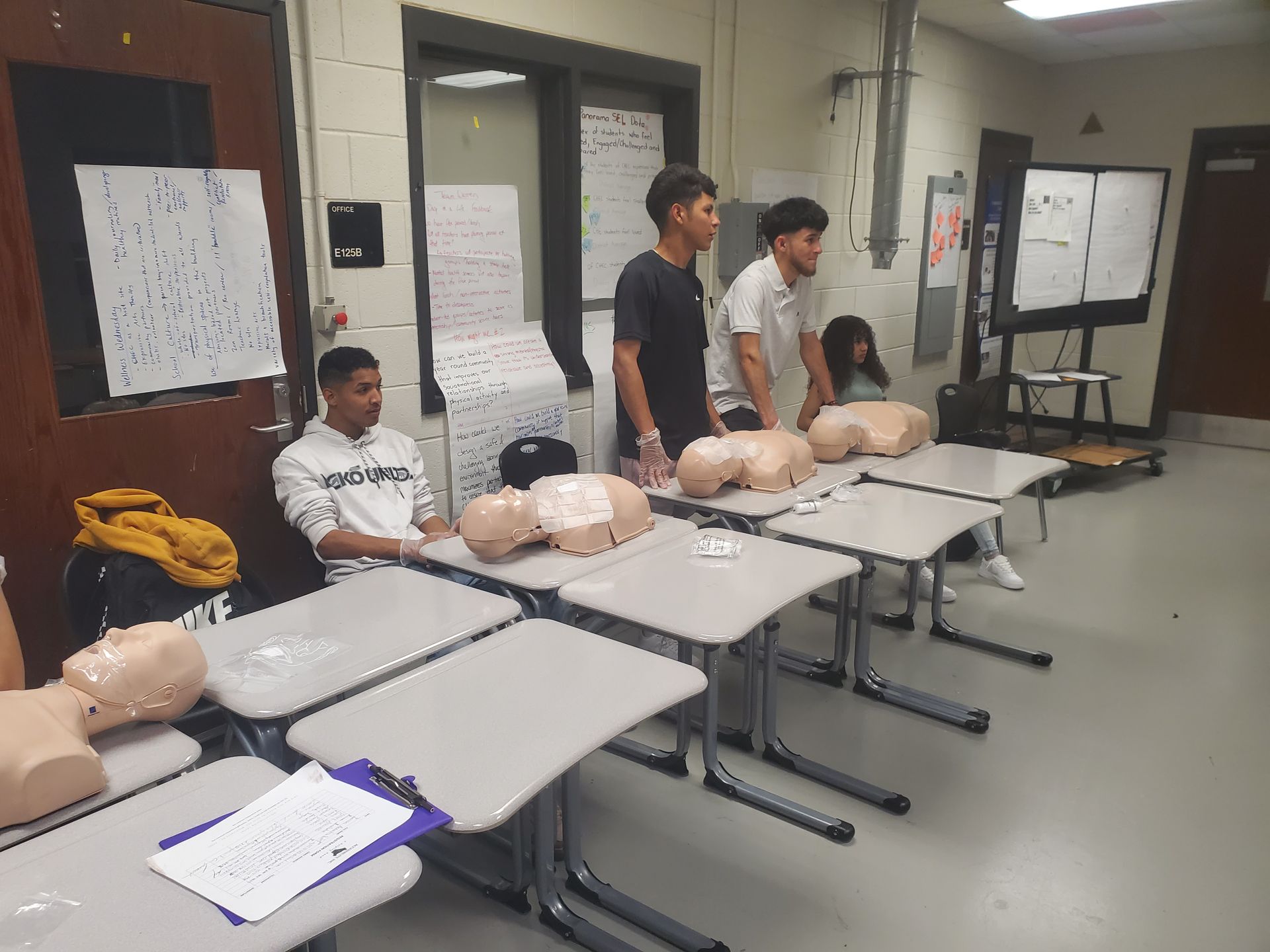 A group of people are sitting at tables in a classroom with mannequins.
