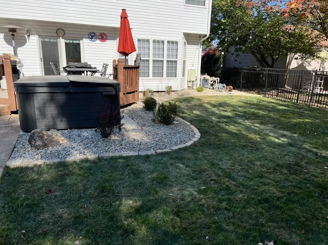 Backyard patio with black hot tub, red umbrella, and stone border beside a white house.