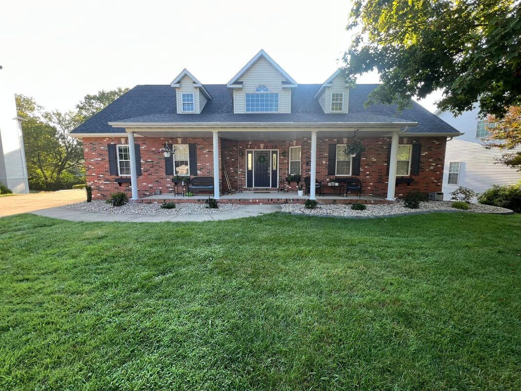 Brick house with a covered front porch, dormer windows, and a green lawn under trees.