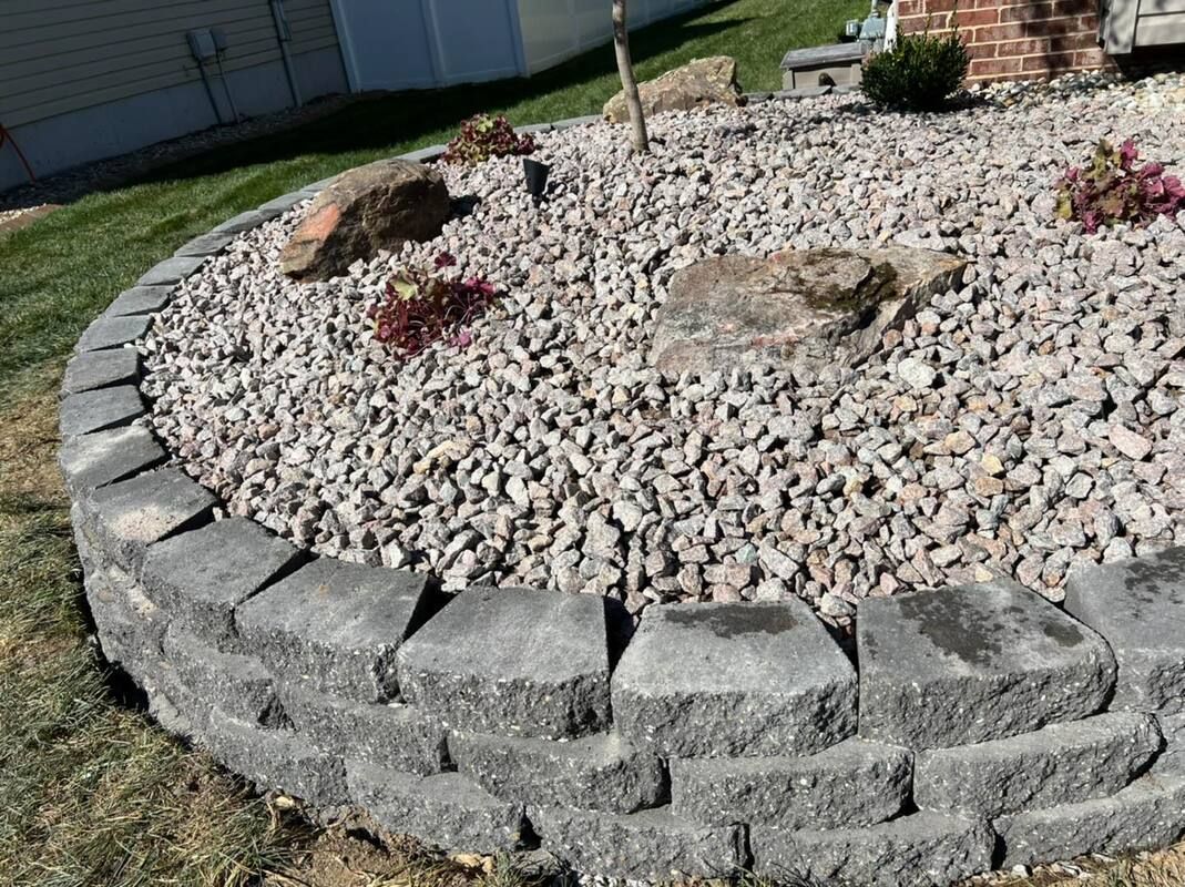 Curved stone retaining wall around a rock garden with small shrubs and a grassy yard
