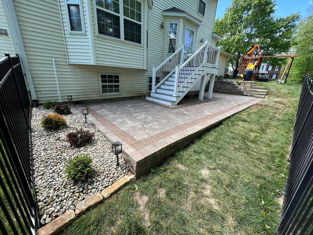 Brick patio beside house stairs and window wells, bordered by gravel and lawn.
