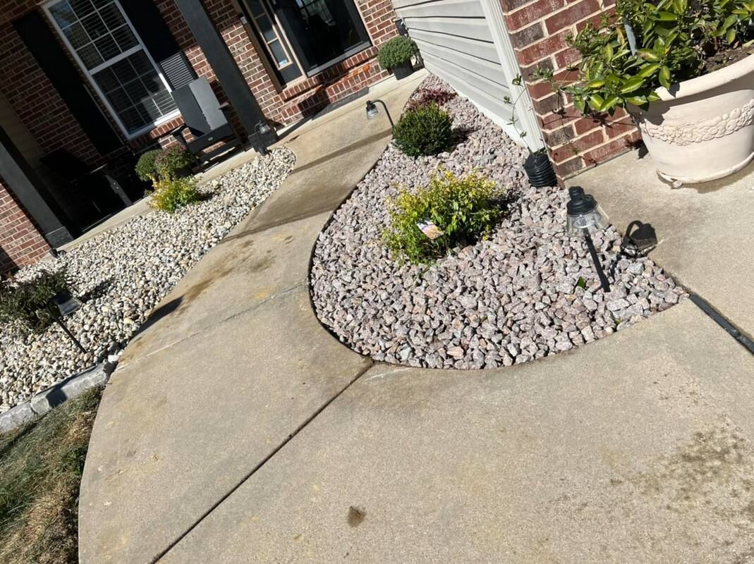 Curved concrete walkway beside a brick building, bordered by gray rock planters with flowers.