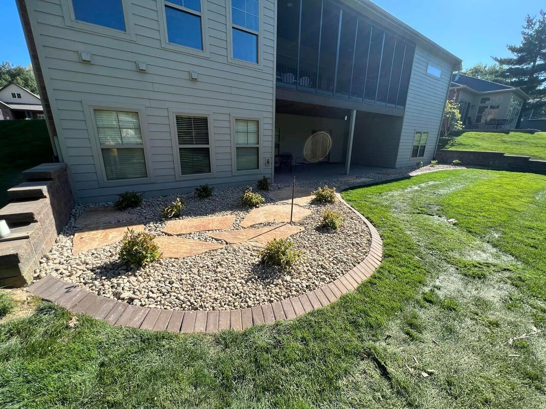 Landscaped front yard with a curved brick flower bed, small shrubs, and a house exterior in sunlight