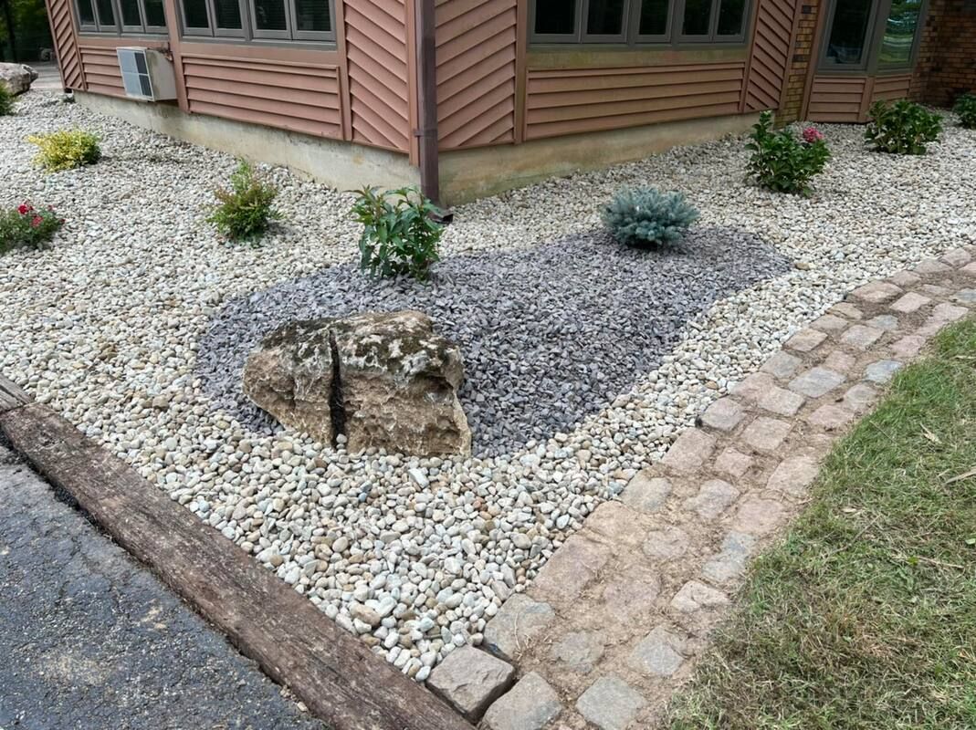 Landscaped rock garden with small shrubs around a tree stump beside a brick house