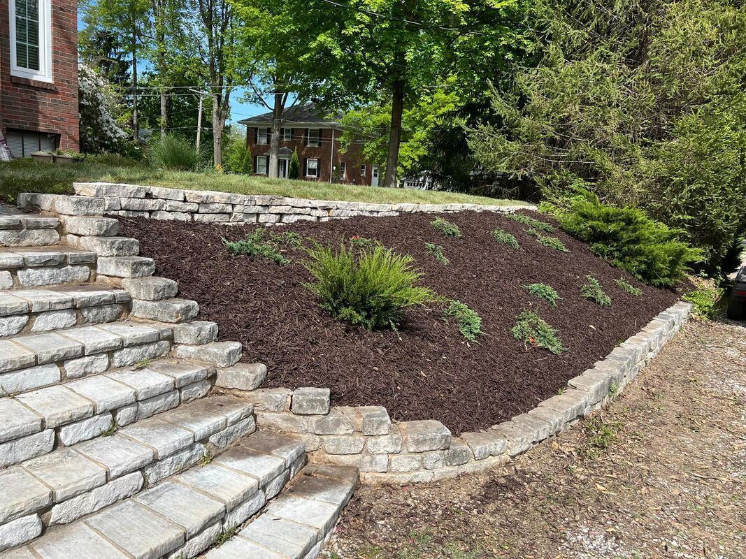 Stone steps beside a mulched hillside with small plants and a house in the background