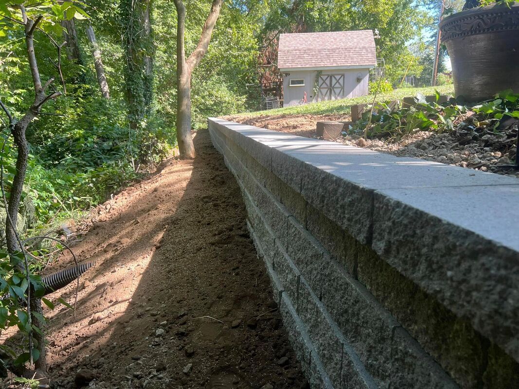 Stone retaining wall beside a dirt path leading to a small house in a wooded area