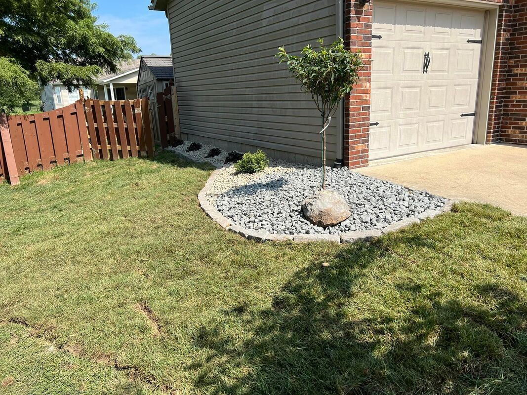 Backyard corner with garage, gravel bed, small shrubs, wooden fence, and lawn in sunlight