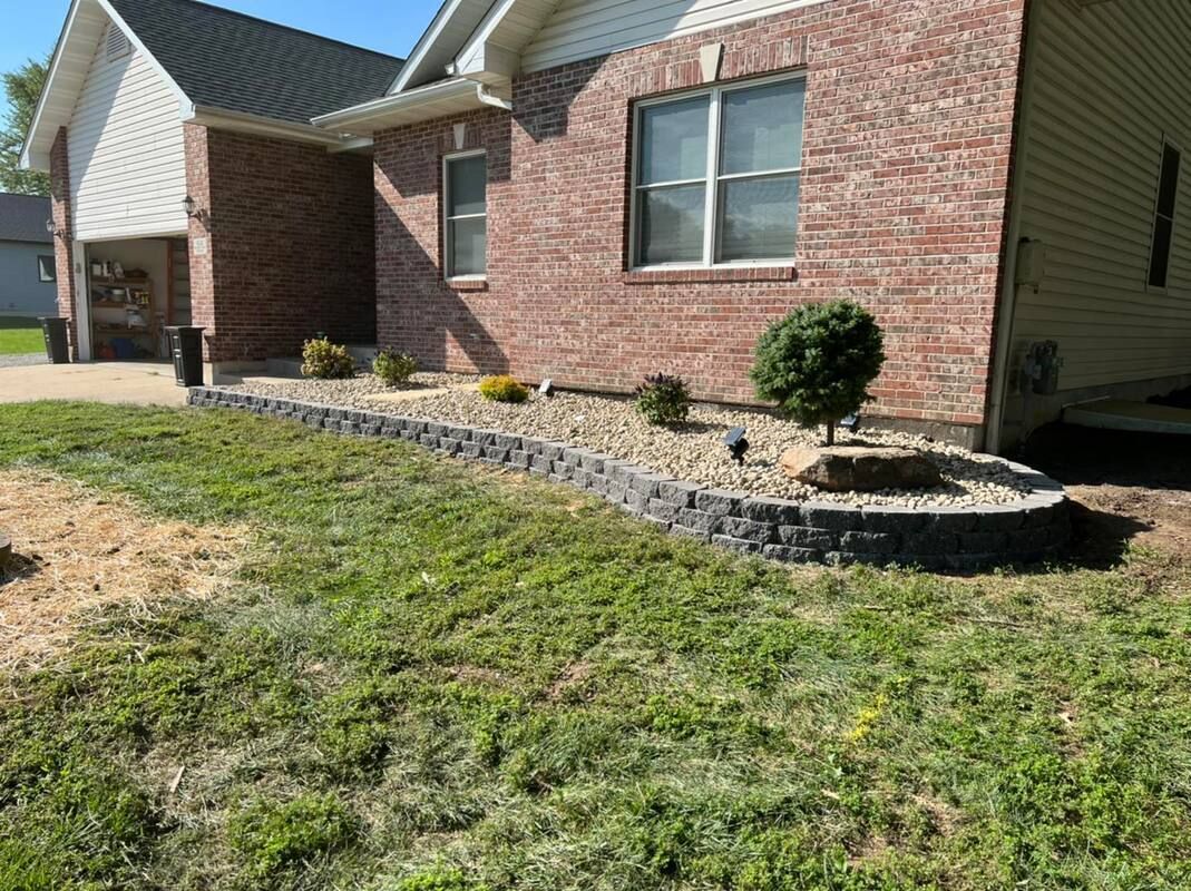 Brick house exterior with a curved stone flower bed and small shrubs beside a grassy yard