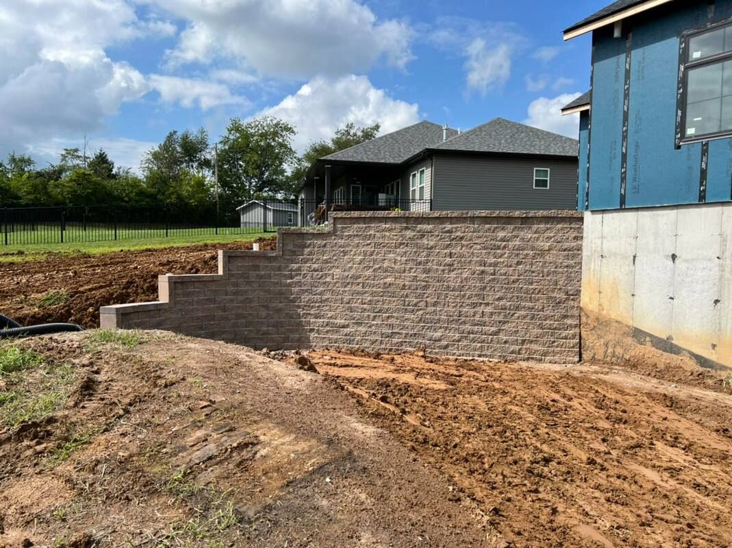 New brick retaining wall beside a muddy construction site and a blue building under a partly cloudy sky