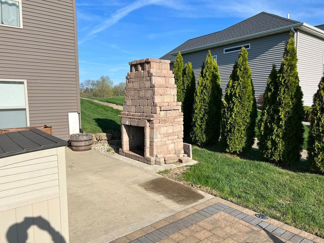 Backyard patio with a brick outdoor fireplace beside a house and tall evergreen shrubs