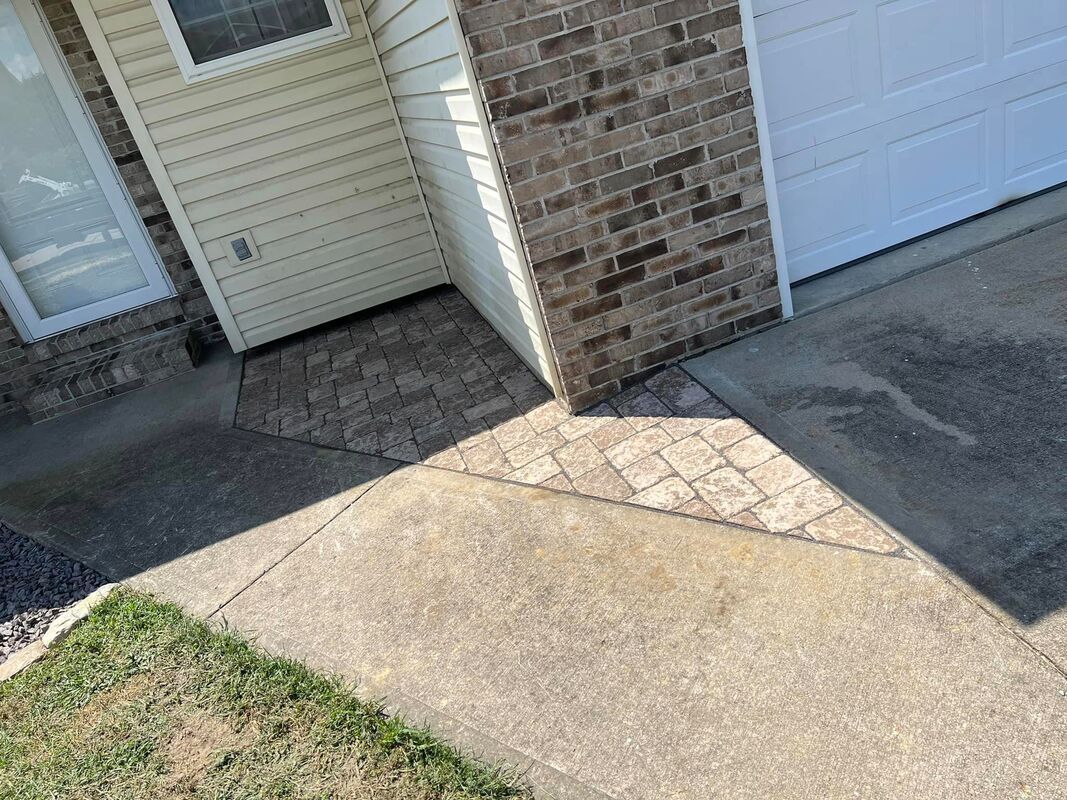 Front entry walkway beside a brick garage, with a concrete path and grass edge in sunlight.