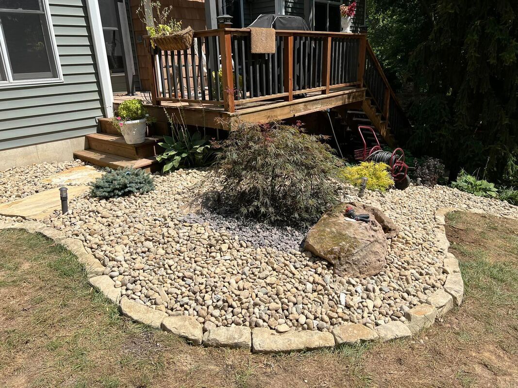 Front yard garden bed with river rocks, shrubs, and a wooden deck beside a house