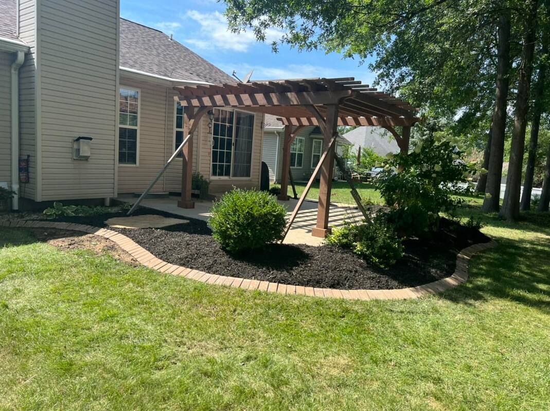 Backyard pergola with wood beams beside a beige house, over a curved mulch bed and green lawn