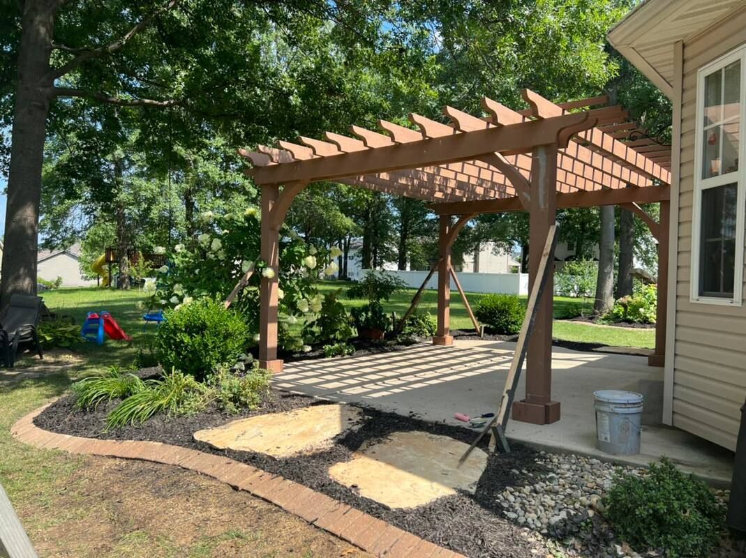 Backyard patio with wooden pergola, stepping stones, mulch beds, and a house exterior.
